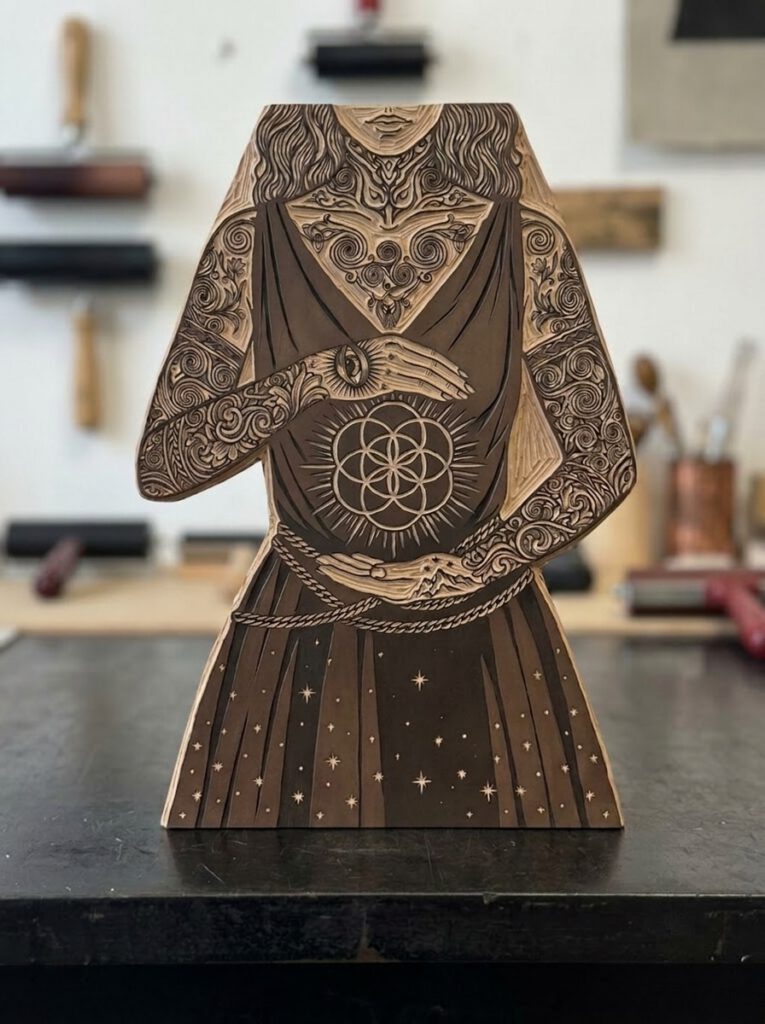A carved wooden linocut relief block prototype depicting Feyre Archeron standing on a printmaking workbench with carving tools and ink rollers blurred in the background.