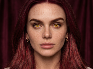 Photorealistic close-up character portrait of Bryce Quinlan featuring natural wine-red hair with dimension, striking golden-amber eyes, and subtle freckles against a dark velvet backdrop.