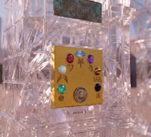 Close-up of the quartz Heart Gate panel in The Old Square of Lunathion, featuring an arched array of seven bezel-set gemstones, engraved district insignias, and a central bronze call button set into a brushed gold plate beneath an aged verdigris plaque.